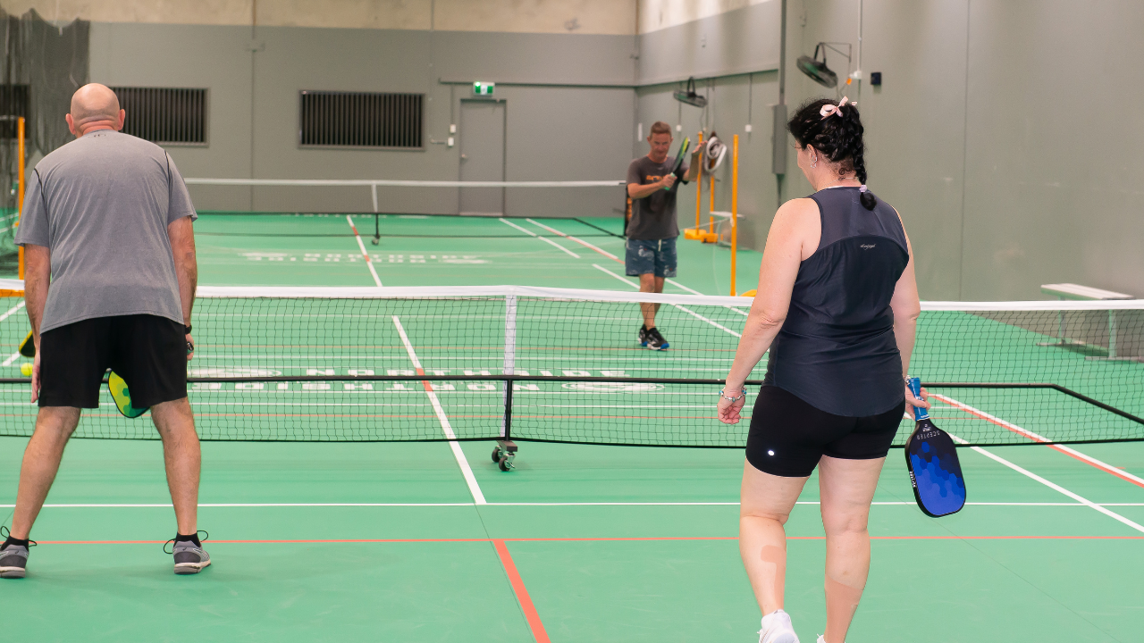 Indoor badminton match at Northside Badminton Centre featuring a male and female player rallying on a blue court inside a modern sports stadium with the Northside Badminton Centre logo displayed on the wall behind them.